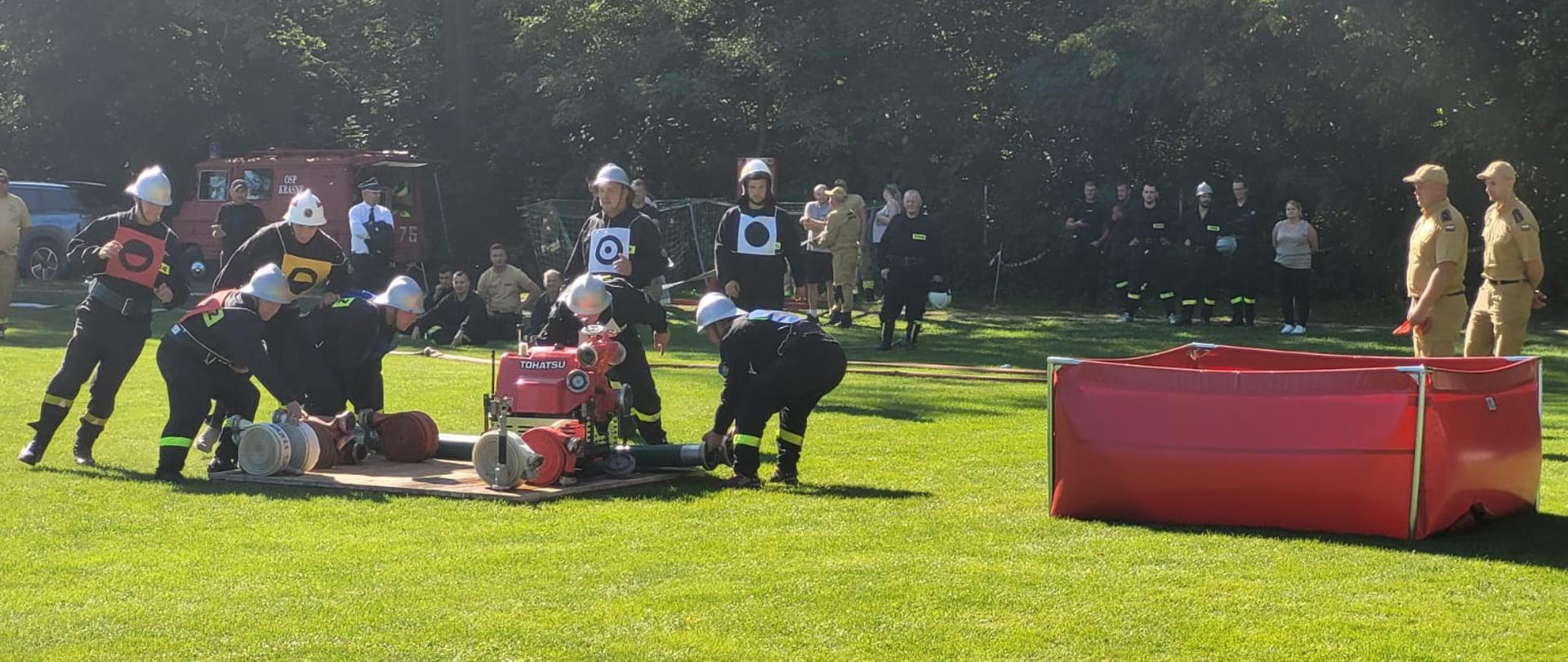 Drużyna OSP wykonuje ćwiczenie bojowe, uruchamiając motopompę i podłączając węże pożarnicze, pod nadzorem sędziów