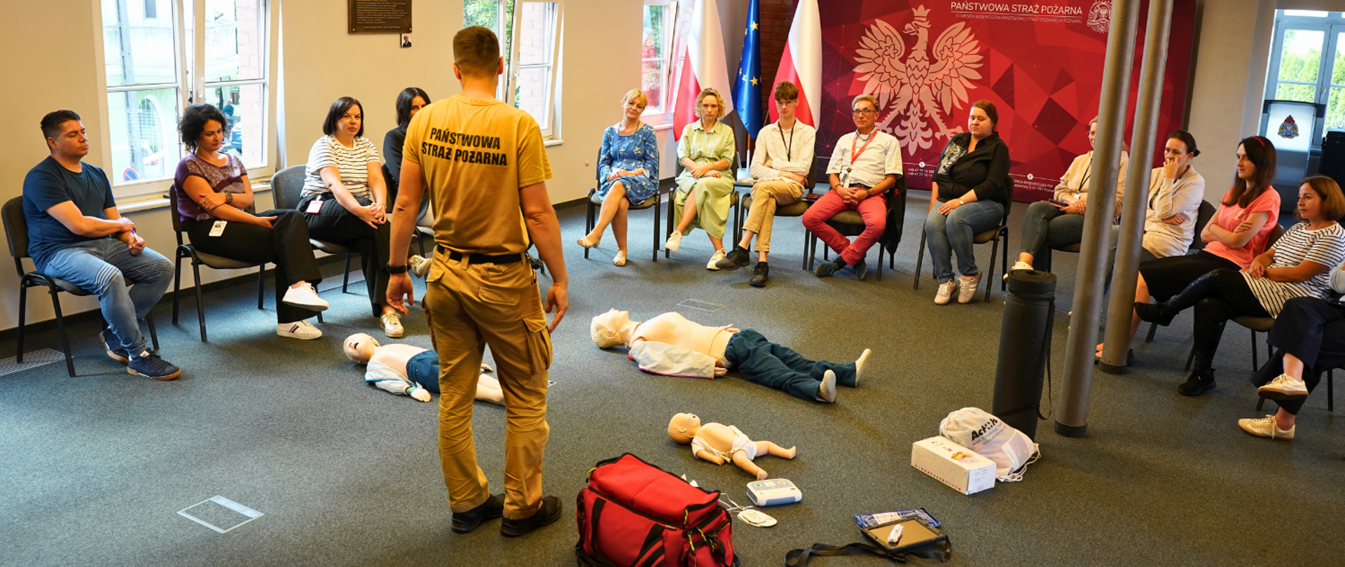 Strażak, który jest ratownikiem medycznym, mówi do wszystkich zebranych na sali, mając przed sobą manekiny. Słuchacze siedzą na krzesłach wokół manekinów, wszyscy są w sali. 