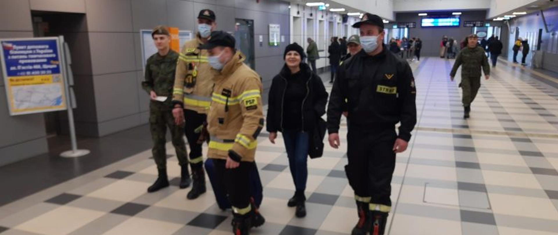 Zdjęcie wykonane na peronie dworca. W centralnym punkcie trzej strażacy oraz kobieta i chłopak w mundurze moro. Cała grupa kieruje się do punktu recepcyjnego. Pani ubrana na czarno i w czapce.
