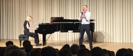 Zdjęcie przedstawia koncert w sali koncertowej. Na scenie mężczyzna śpiewa do mikrofonu, stojąc przy statywie, ubrany w białą koszulę i ciemne spodnie. Obok niego przy fortepianie koncertowym siedzi pianistka w czarnej sukni, akompaniująca wokaliście. Scena oświetlona jest ciepłym światłem, w tle widoczna jest jasna, pionowa okładzina ściany