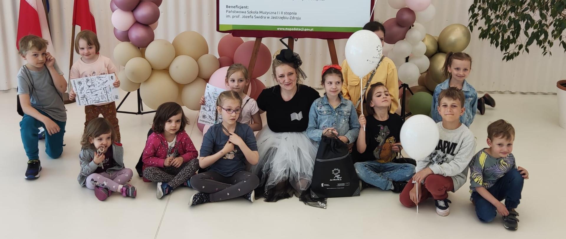 Kobieta ubrana w tiulową białą spódniczkę , czarną bluzkę z logo sali koncertowej i czarny kapelusik z nutką siedzi na białej podłodze z jedenaściorgiem dzieci, za nimi tablica informująca o dotacji środków z UE na rozbudowę budynku szkoły o salę koncertową. Tablica jest udekorowaną dookoła balonami.