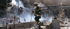 Zdjęcie przedstawia gruzowisko po zawaleniu się dwóch wież World Trade Center. Z gruzów unosi się dym. Wśród gruzów, zawalonych elementów konstrukcyjnych budynków widać strażaków – ratowników, którzy przeszukują miejsce katastrofy w poszukiwaniu osób ocalałych, które mogły przeżyć zamach terrorystyczny. 