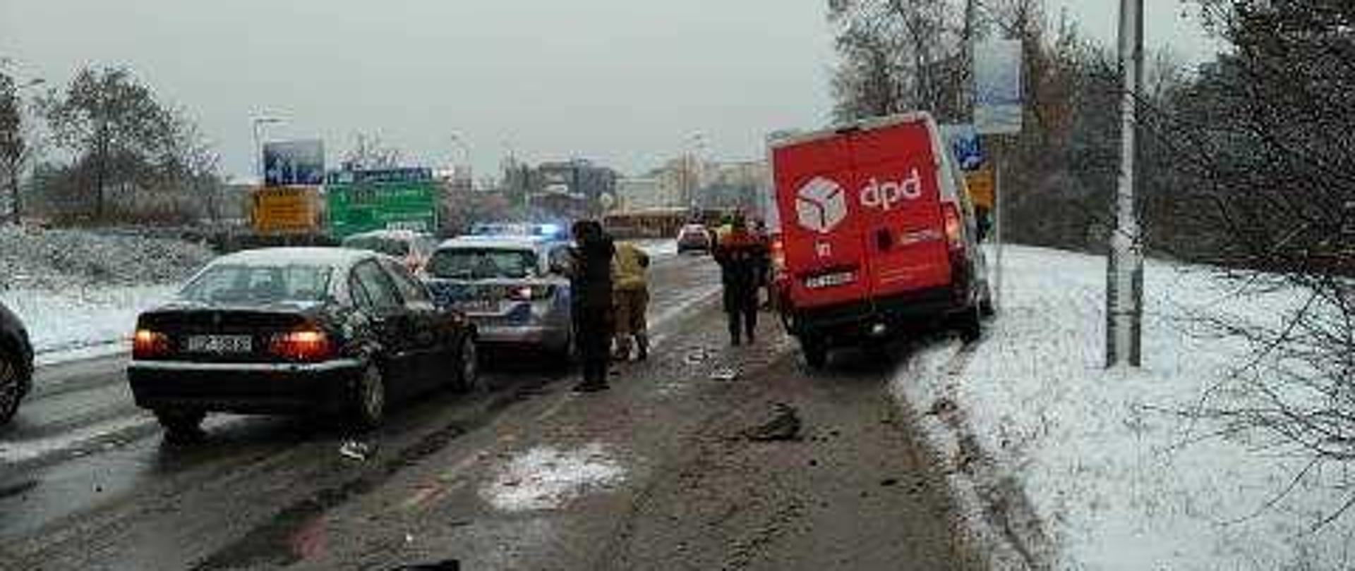 Zdjęcie przedstawia ulicę Tarnowską w Kielcach. Po lewej stronie, stoją dwa samochody biorące udział w zdarzeniu. Przy nich stoją osoby postronne oraz strażak. Po prawej samochód dostawczy również biorący udział w wypadku.