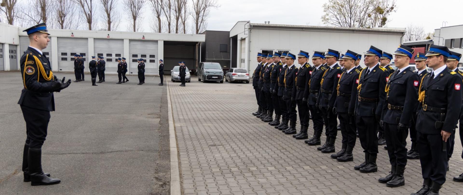 Kompania reprezentacyjna PSP stoi w kilku równych szeregach na placu przed budynkiem, a dowódca prowadzi odprawę.