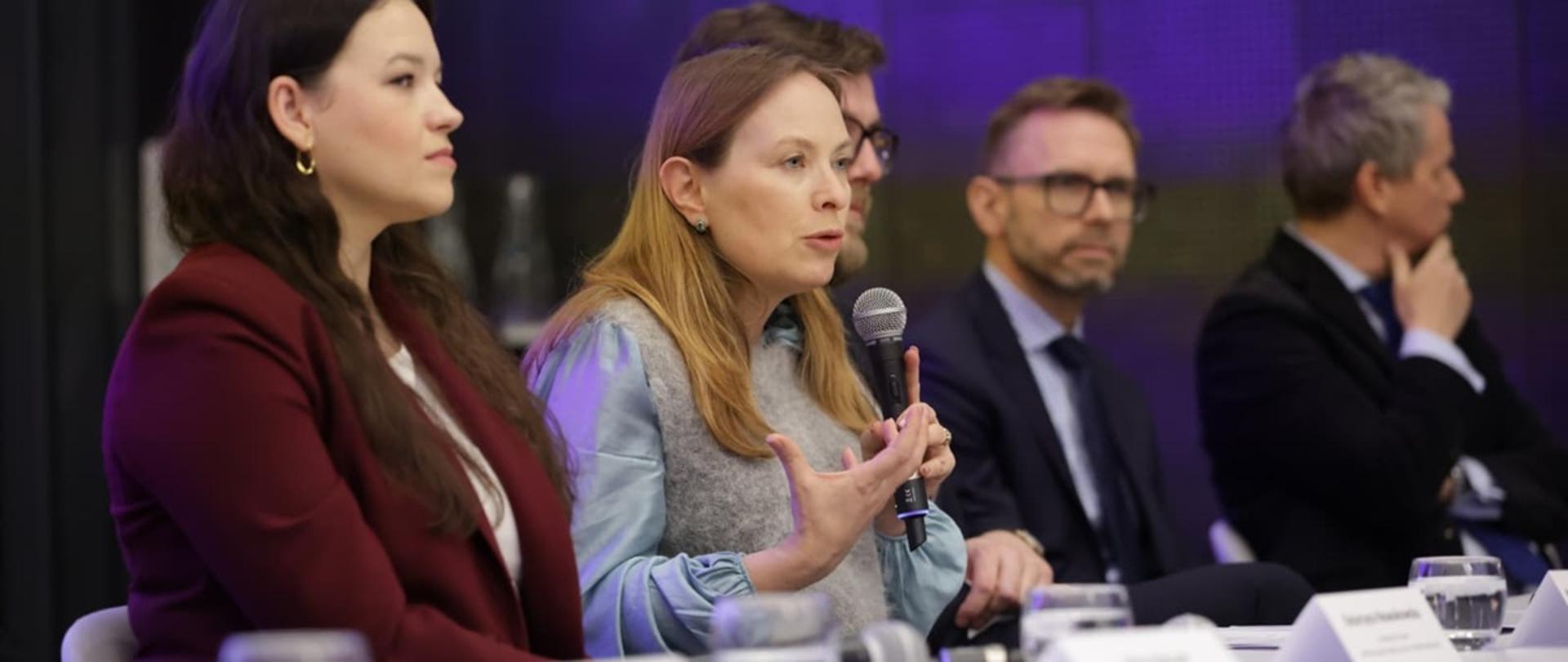 Minister funduszy i polityki regionalnej Katarzyna Pełczyńska-Nałęcz siedzi przy stole i przemawia do mikrofonu trzymanego w dłoniach. Po jej lewej stronie siedzi wicedyrektor Departamentu Koordynacji Krajowego Planu Odbudowy w MFiPR Mateusz Gaczyński.