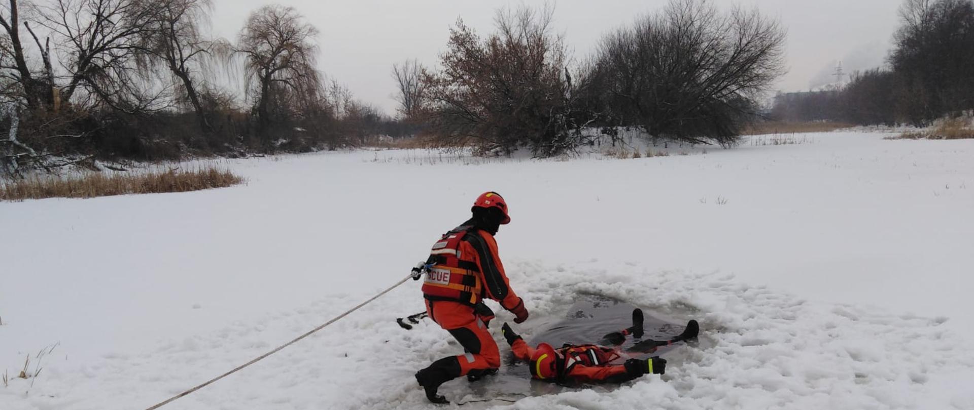 Dwóch strażaków na śniegu. Jeden leży w wodzie. W tle śnieg i drzewa. 