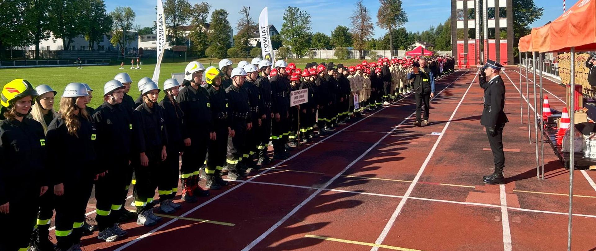 Długi rząd młodzieży w czarnych mundurach strażackich z białymi i czerwonymi hełmami, stojącej w szyku na bieżni stadionu. Po prawej stronie strażak w mundurze galowym salutuje. W tle wieża treningowa do ćwiczeń wysokościowych z numeracją "1 2 3 4", flaga Polski oraz zielone boisko z drzewami.