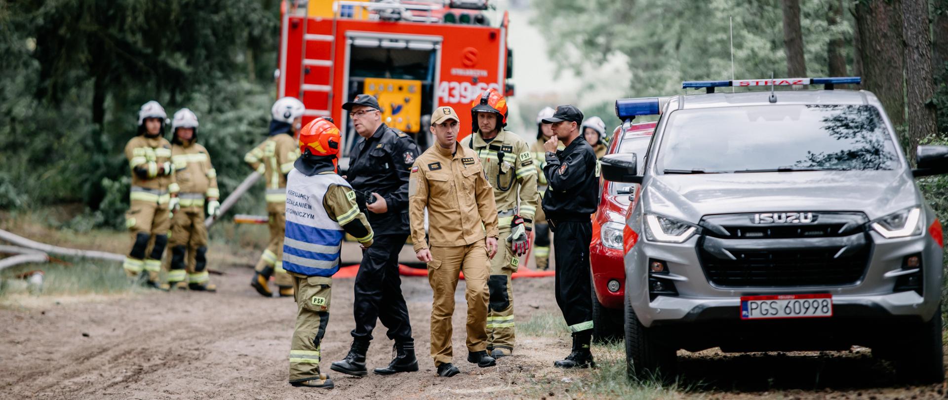 Zdjęcie przedstawia strażaków oraz druhów podczas ćwiczeń. Stoją oni i rozmawiają ze sobą. W tle wóz strażacki. Z prawej strony dwa samochody SLRR.
