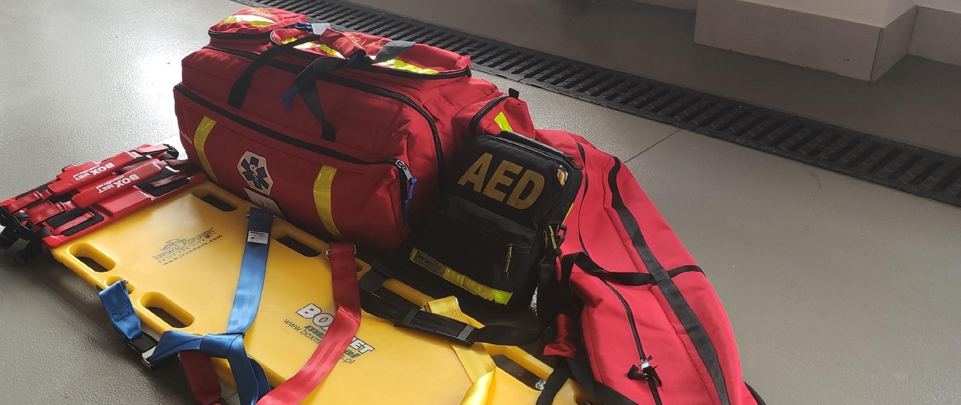 Zdjęcie obrazuje nosze typu deska ortopedyczna walizkę medyczną, szyny do stabilizacji,AED