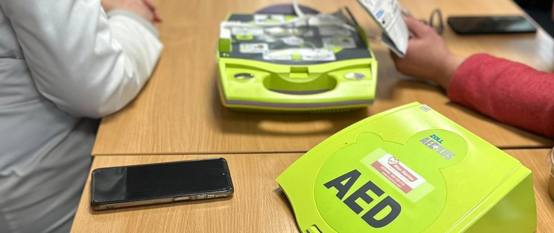 Na fotografii widać zielone urządzenie AED (automatyczny defibrylator zewnętrzny) umieszczone na drewnianym stole. W tle znajduje się otwarte etui z elementami defibrylatora, w tym elektrodami. 