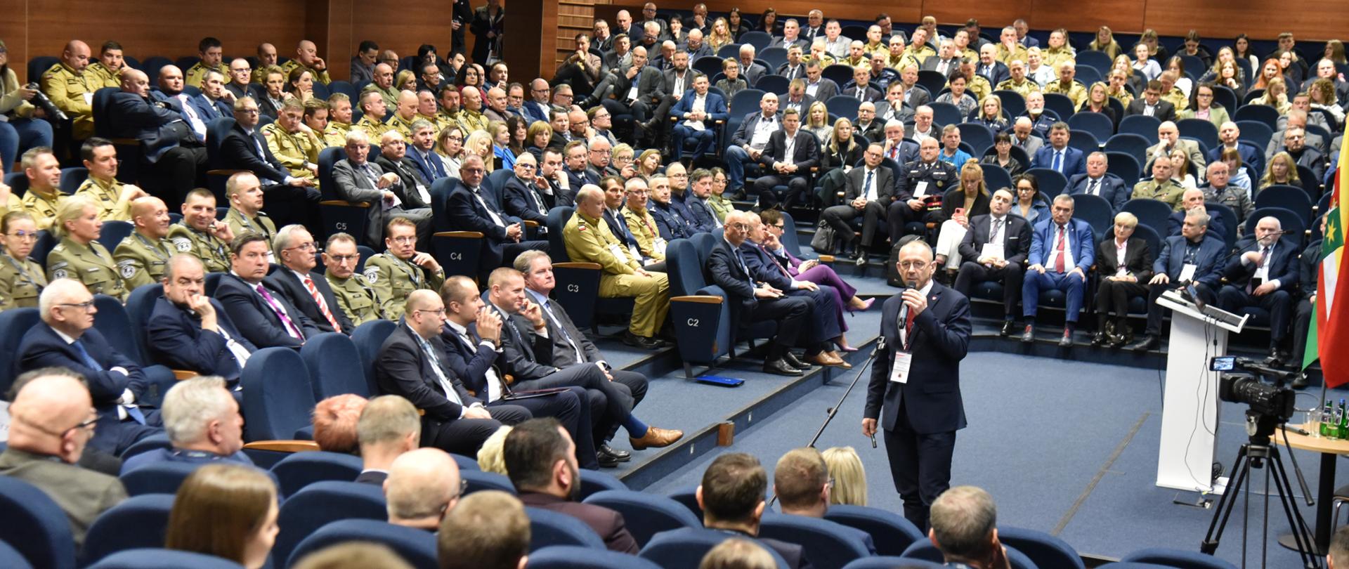Grupa osób siedzących na dużej sali, są wśród funkcjonariusze Policji i Państwowej Straży Pożarnej, Straży Granicznej. Pośrodku mężczyzna z mikrofonem. 