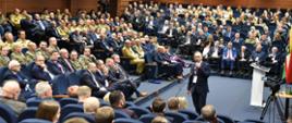Grupa osób siedzących na dużej sali, są wśród funkcjonariusze Policji i Państwowej Straży Pożarnej, Straży Granicznej. Pośrodku mężczyzna z mikrofonem. 