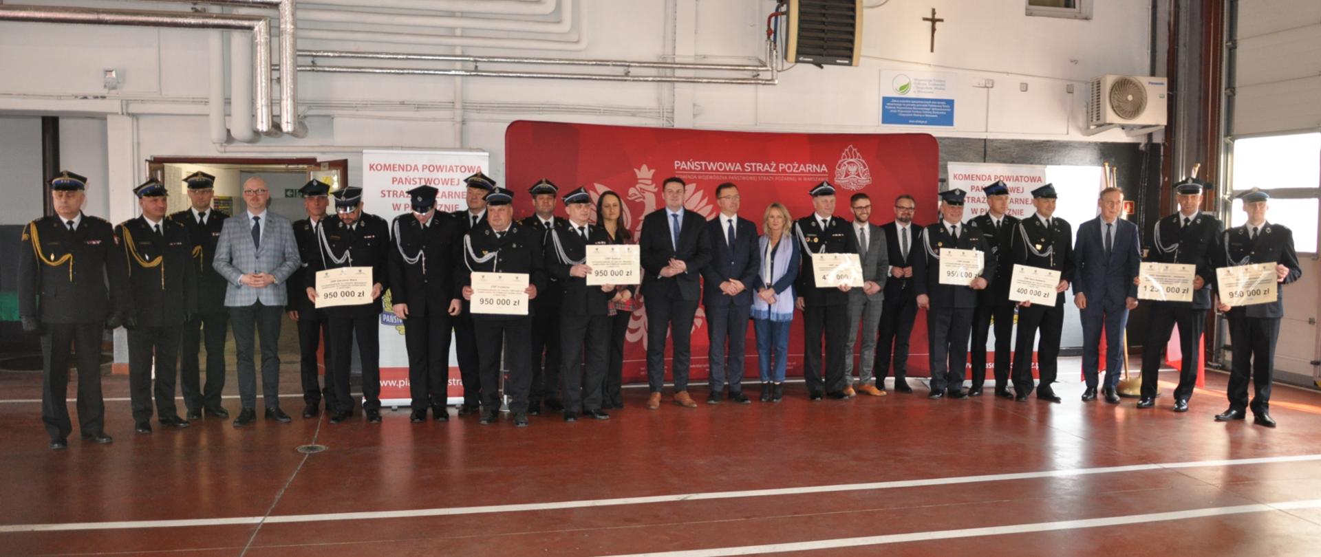 Zdjęcie przedstawia ustawionych na tle baneru strażaków oraz polityków. Strażacy trzymają w rękach promesy na zakup samochodów pożarniczych.