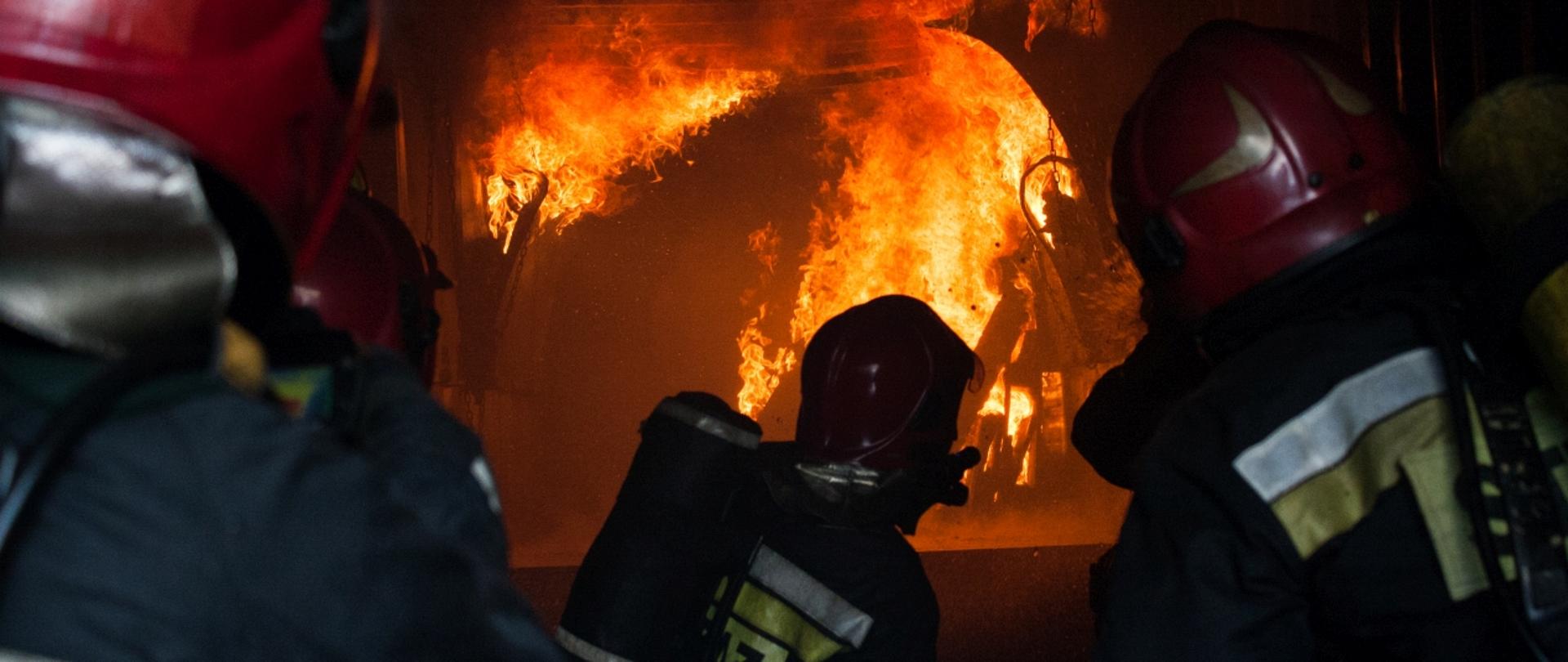Zdjęcie poglądowe przedstawia strażaków w umundurowaniu bojowym w aparatach ochrony układu oddechowego. Strażacy znajdują się w komorze rozgorzeniowej. 