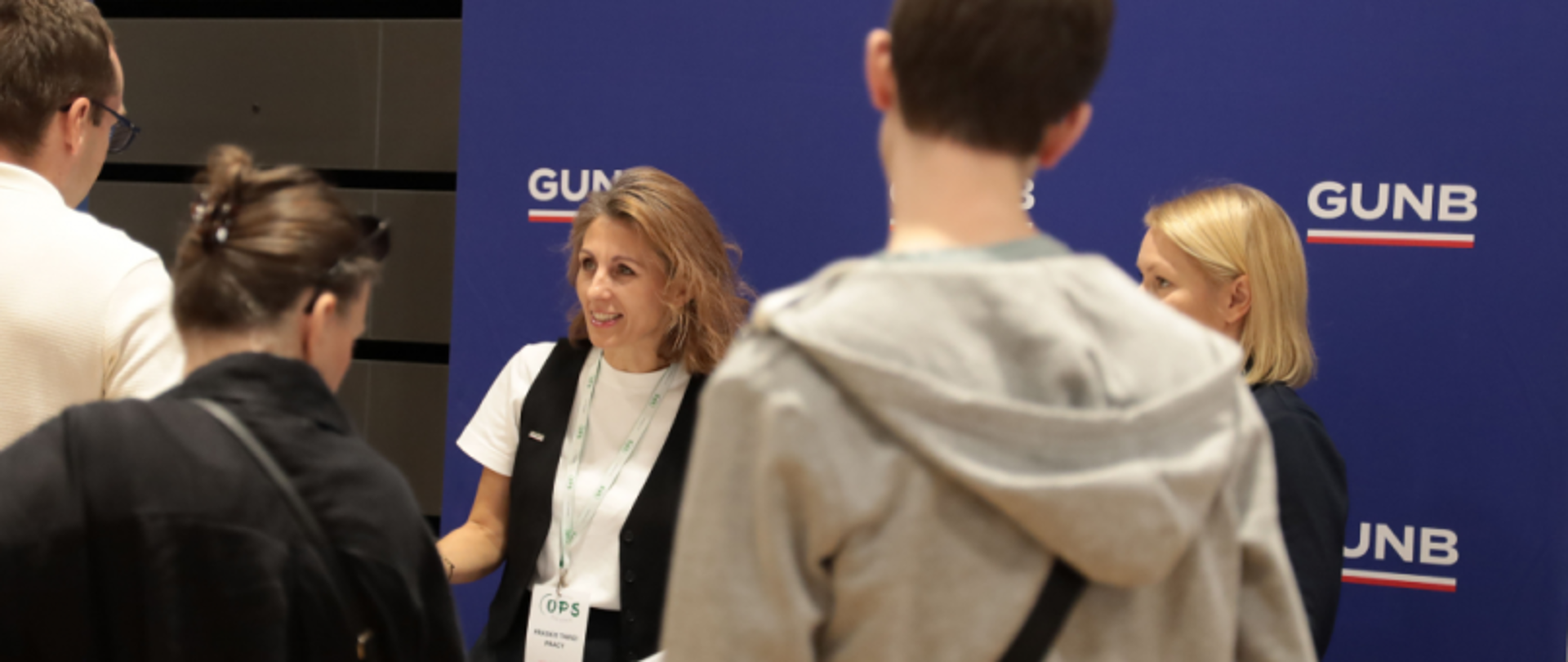 Uczestnicy wydarzenia na tle ścianki „GUNB”, niektórzy z identyfikatorami, w trakcie rozmowy.