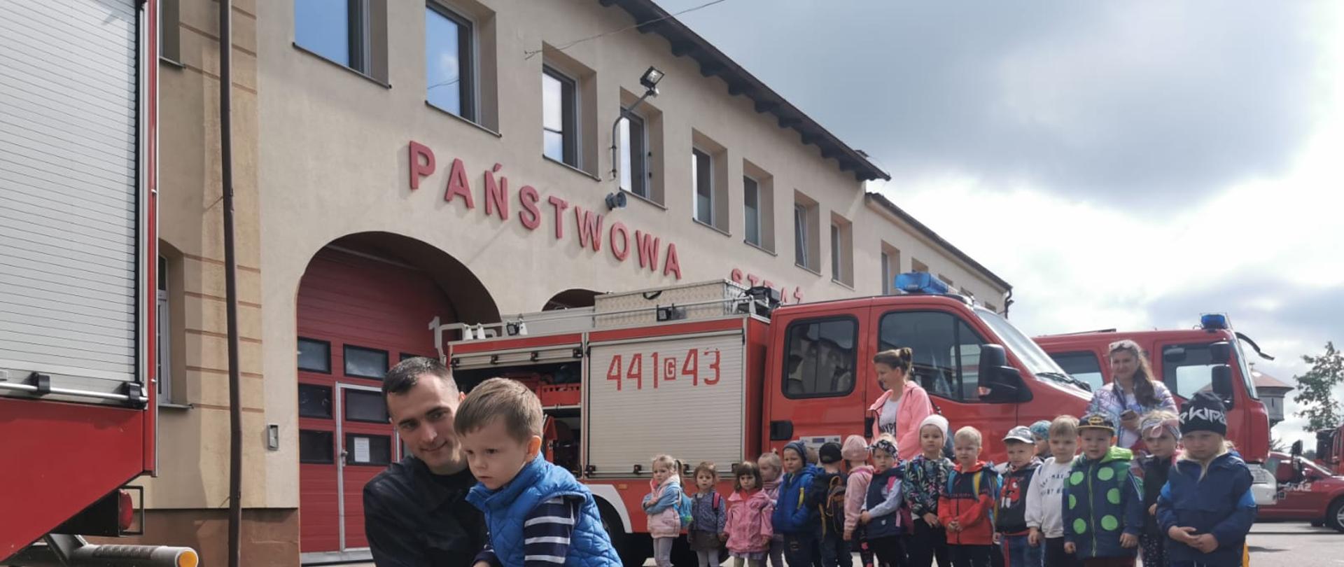 Na fotografii strażak pokazuje chłopcowi w jaki sposób trzymać wąż gaśniczy, w tle wozy gaśnicze oraz dzieci.