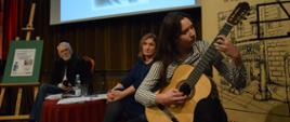 Gitarzystka podczas występu, w tle prowadząca spotkanie poetyckie oraz literat. 