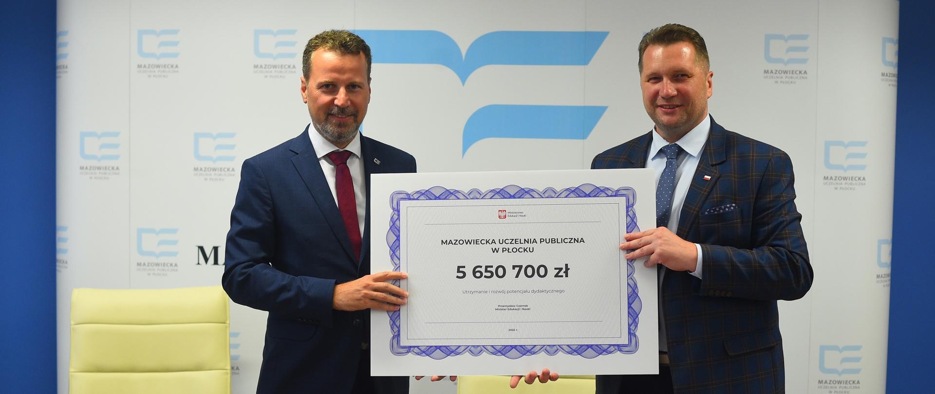 Minister stoi za stołem i prezentuje symboliczny czek na kwotę 5 650 700 zł.