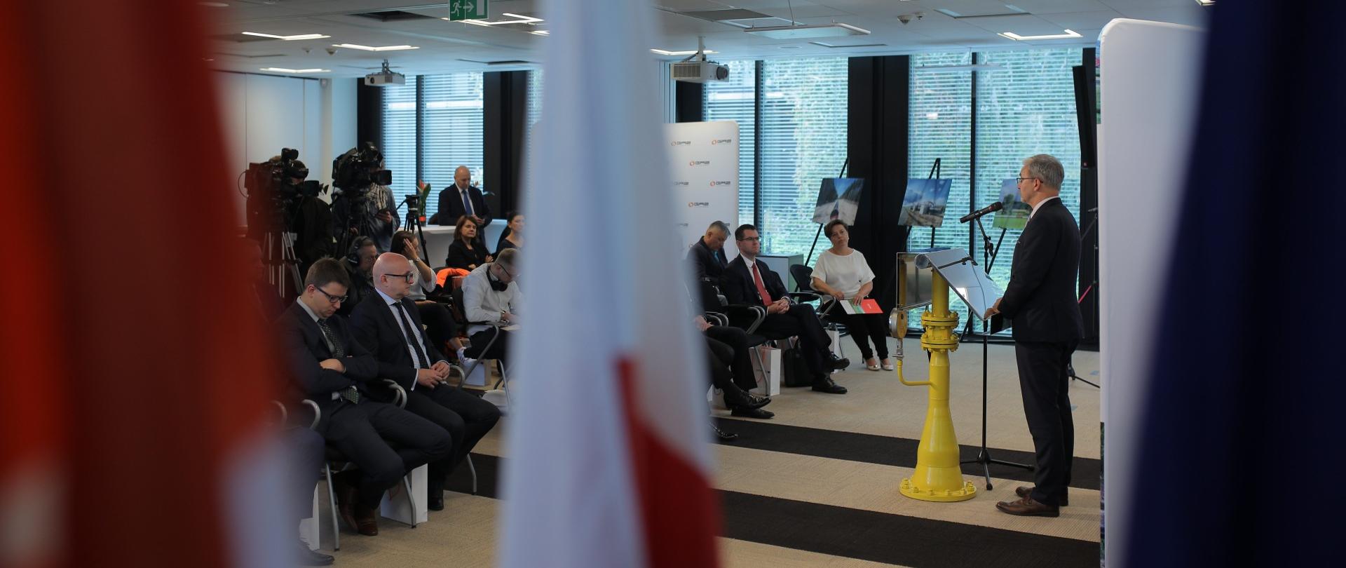 Minister Kwieciński stoi przy mównicy. Na przeciwko siedzą pozostali uczestnicy spotkania, z tylu sali stoją kamery. 