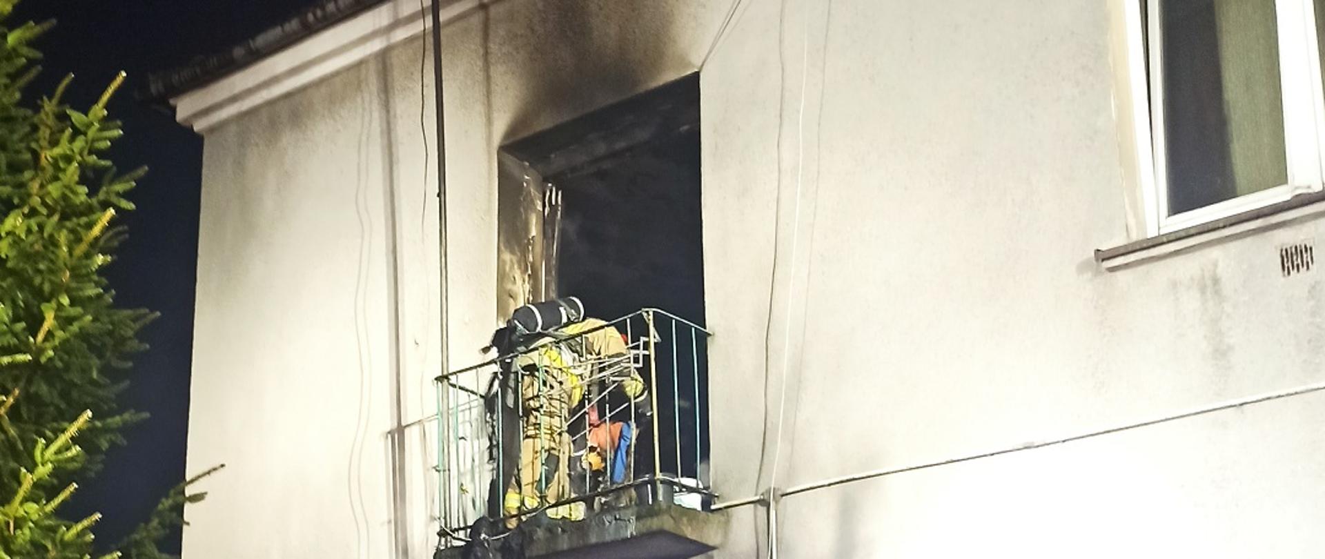 Zdjęcie obrazuje strażaka pracującego na balkonie budynku wielokondygnacyjnego na ścianie widoczne oznaki pożaru