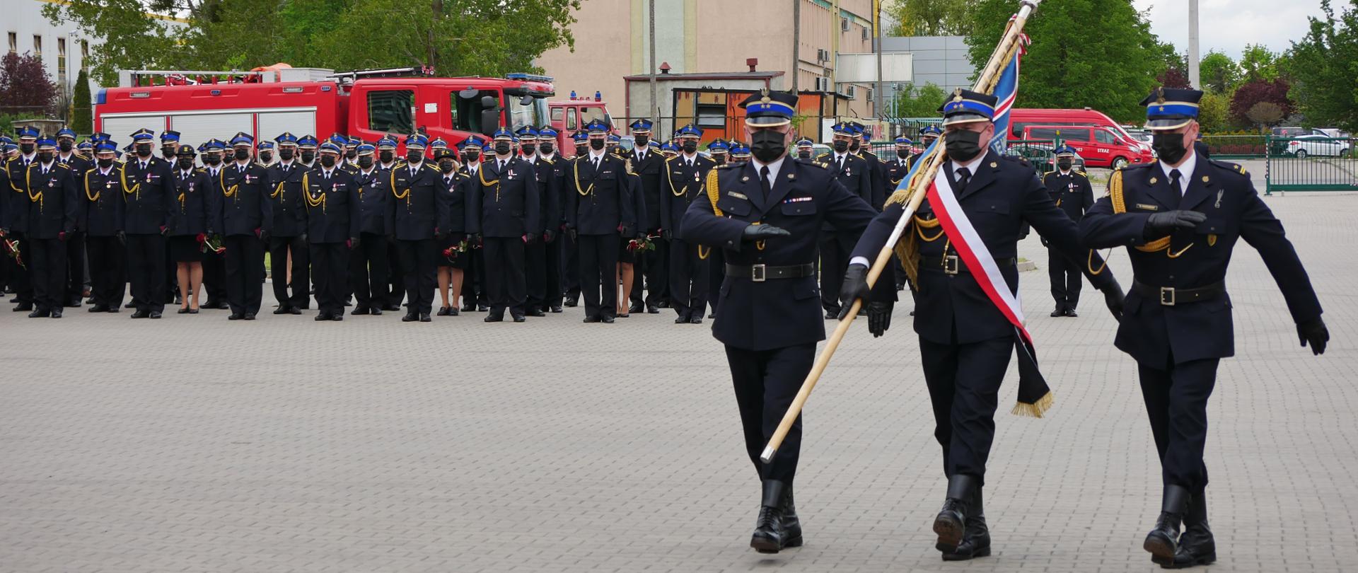 Plac jednostki ratowniczo gaśniczej w Gorzowie Wielkopolskim. Przed jednostką odbywa się uroczystość podczas której wyróżnienia, awansowani i mianowani strażacy z województwa lubuskiego ustawieni są w szeregach. na pierwszym planie z prawej strony maszeruje poczet sztandarowy złożony z trzech strażaków.