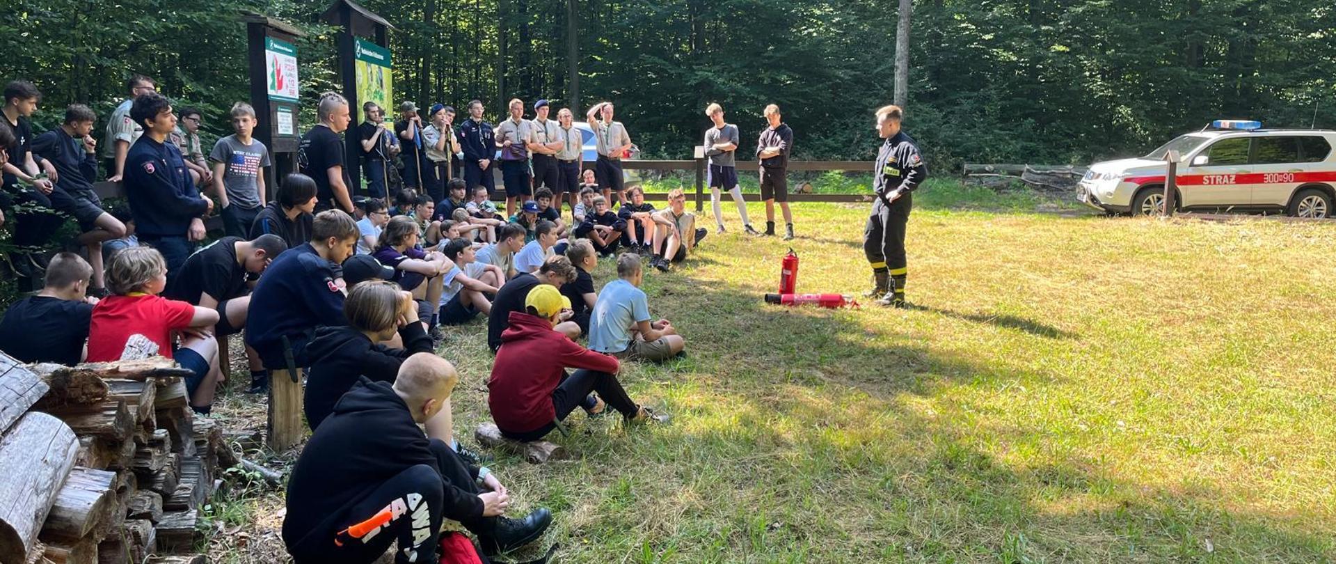 Zdjęcie przedstawia szkolenie z zakresu użycia podręcznego sprzętu gaśniczego. Uczestnicy obozu - dzieci słuchają szkolenia strażaka przed którym ustawione są dwie gaśnice. W tle widoczny pojazd Państwowej Straży Pożarnej.