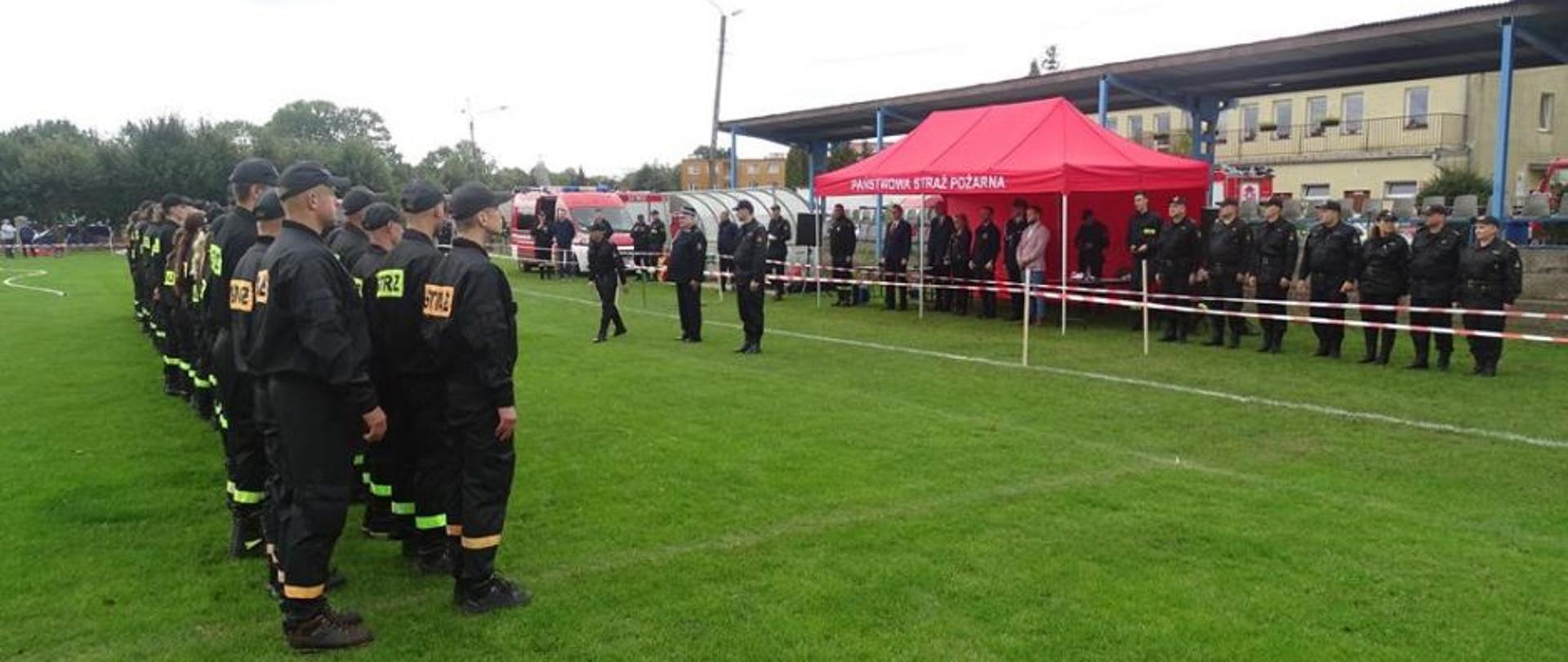 Zbiórka strażaków na boisku przed zawodami. Po lewej stronie stoją w dwóch rzędach strażacy biorący udział w zawodach, na przeciwko nich stroją organizatorzy i zaproszeni goście.