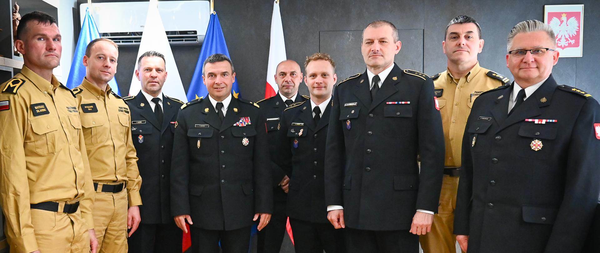 Pamiątkowe zdjęcie grupowe dziewięciu strażaków stojących w dwóch rzędach w gabinecie. Większość w ciemnych garniturach wyjściowych, kilku w piaskowych mundurach służbowych. Po lewej stronie szafka z pucharami i medalami, w tle flagi i godło na ścianie.