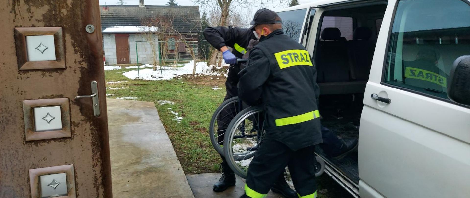 Zdjęcie przedstawia dwóch strażaków przenoszących pomiędzy domem a pojazdem na wózku osobę niepełnosprawną. Z lewej strony znajduje się budynek mieszkalny z otwartymi drzwiami, z prawej strony zaś pojazd przystosowany do transportu osób niepełnosprawnych. 