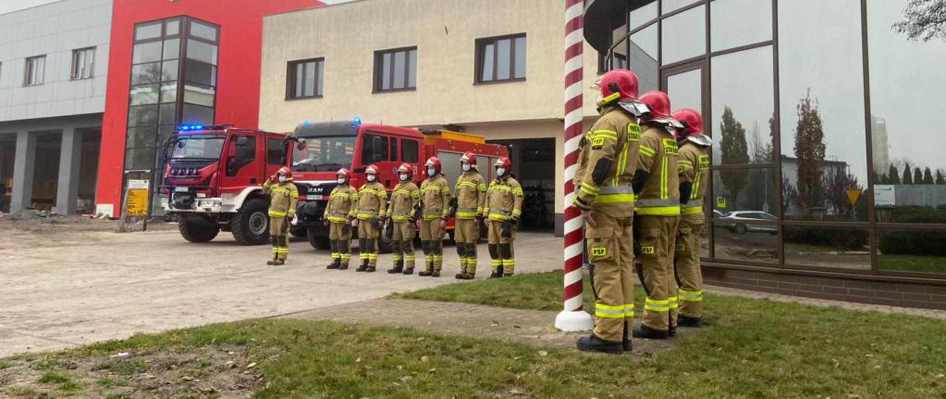 Na zdjęciu widnieją strażacy Państwowej Straży Pożarnej w Pile w trakcie uroczystego wciągnięcia flagi na maszt i odśpiewania hymnu państwowego. Jest dzień, pochmurno, bez opadów. 
