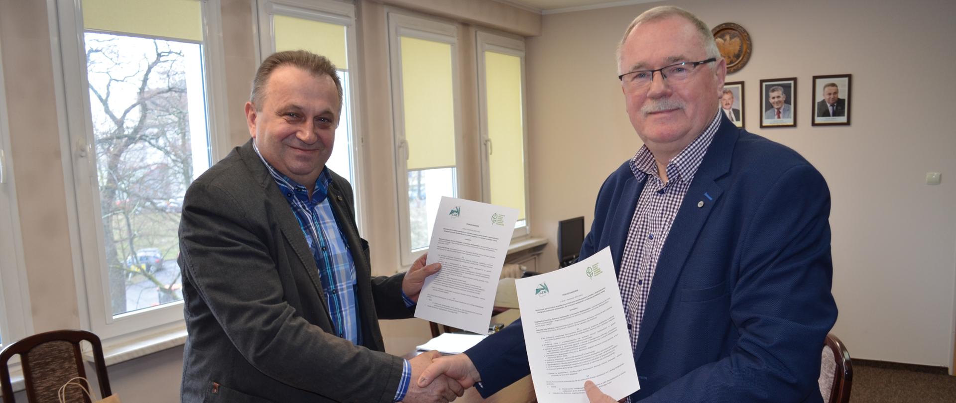 Dwóch mężczyzn ubranych w marynarki, pozuje do zdjęcia, ściskając sobie dłonie. W drugiej ręce każdy z nich trzyma białą zapisaną treścią porozumienia.