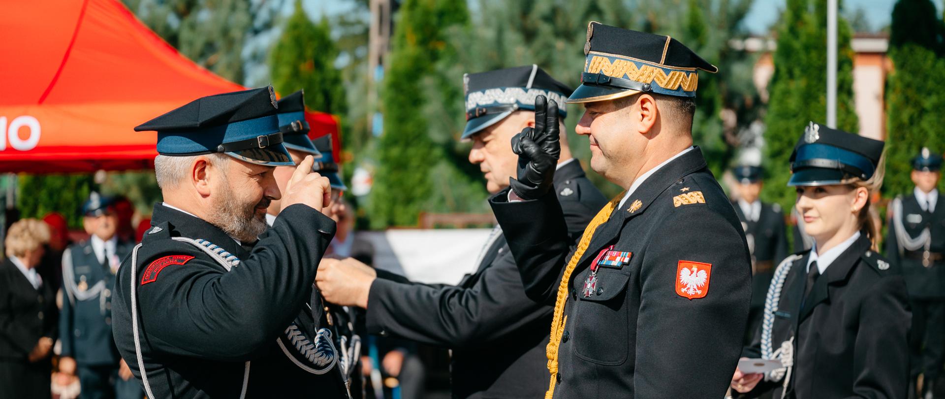Zbliżenie z profilu przedstawia moment nakładania odznaczenia na mundur wyższego rangą strażaka przez Mazowieckiego Komendanta Wojewódzkiego PSP nadbryg. Artura Gonerę. Strażacy ubrani są w galowe mundury z orłem białym na czerwonym tle naszytym na rękawie oraz bogatymi dystynkcjami na pagonach. W tle widoczny jest fragment czerwonego namiotu oraz zielone drzewa. Pogoda jest słoneczna i jasna. Zdjęcie cechuje się dużą podniosłością, skupieniem i dostojeństwem.