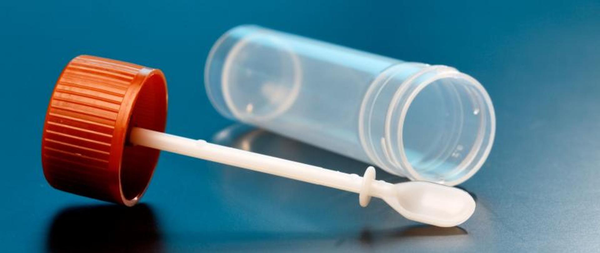 Grafika przewdstawia plastikowy pojemnik do poboru próbki kału do badań na nosicielstwo z łopatką i czerwoną zakrętką