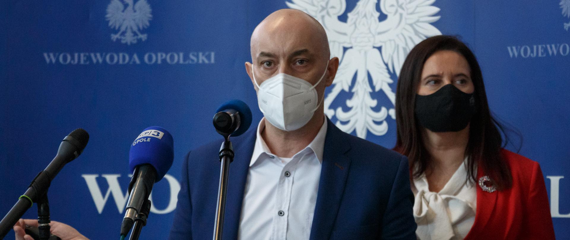 Były już wojewoda opolski Adrian Czubak przemawia do zgromadzonych dziennikarzy. Stoi na ściance granatowej z napisem wojewoda opolski. Stoi przed szklaną mównicą, ma otwarte dłonie. W jednej z nich trzyma pilot do prezentacji. Obok niego stoi poseł Porowska, ubrana w czerwoną garsonkę. Poseł spogląda w bok. 