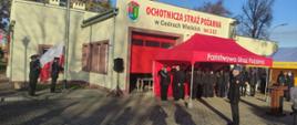 Trzech strażaków ochotniczej straży pożarnej wciąga flagę Polski na maszt strażak stoi na baczność oddając honor trzech pozostali strażacy stoją pod namiotem oddając honory namiot z napisem Państwowa Straż Pożarna ustawiony jest przed budynkiem.