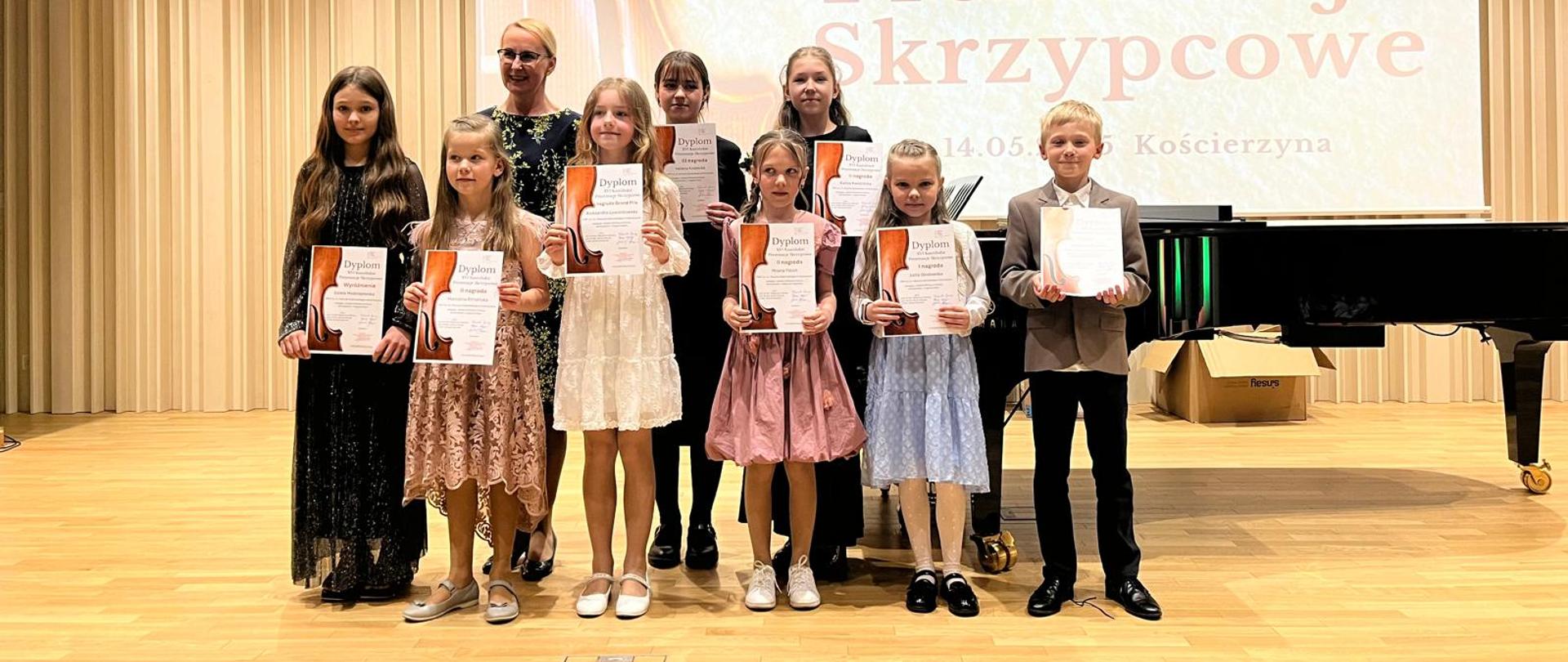 Na scenie na drewnianej podłodze stoi grupa uczniów. Wszyscy trzymają dyplom. Za nimi kobieta blondynka w okularach w granatowej sukience w kwiaty. W tle grafika skrzypiec i napis "XVI Kaszubskie Prezentacje Skrzypcowe". Tło obrazka w odcieniach beżu.