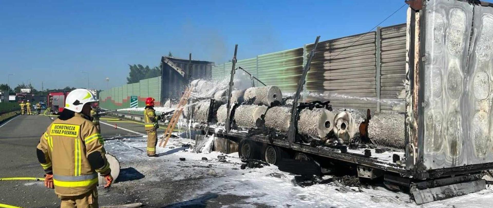 Na drodze ekspresowej S8 stoi spalona naczepa samochodu ciężarowego z pozostałościami folii w rolkach, która była w niej przewożona. Dwóch strażaków dogasza naczepę. 