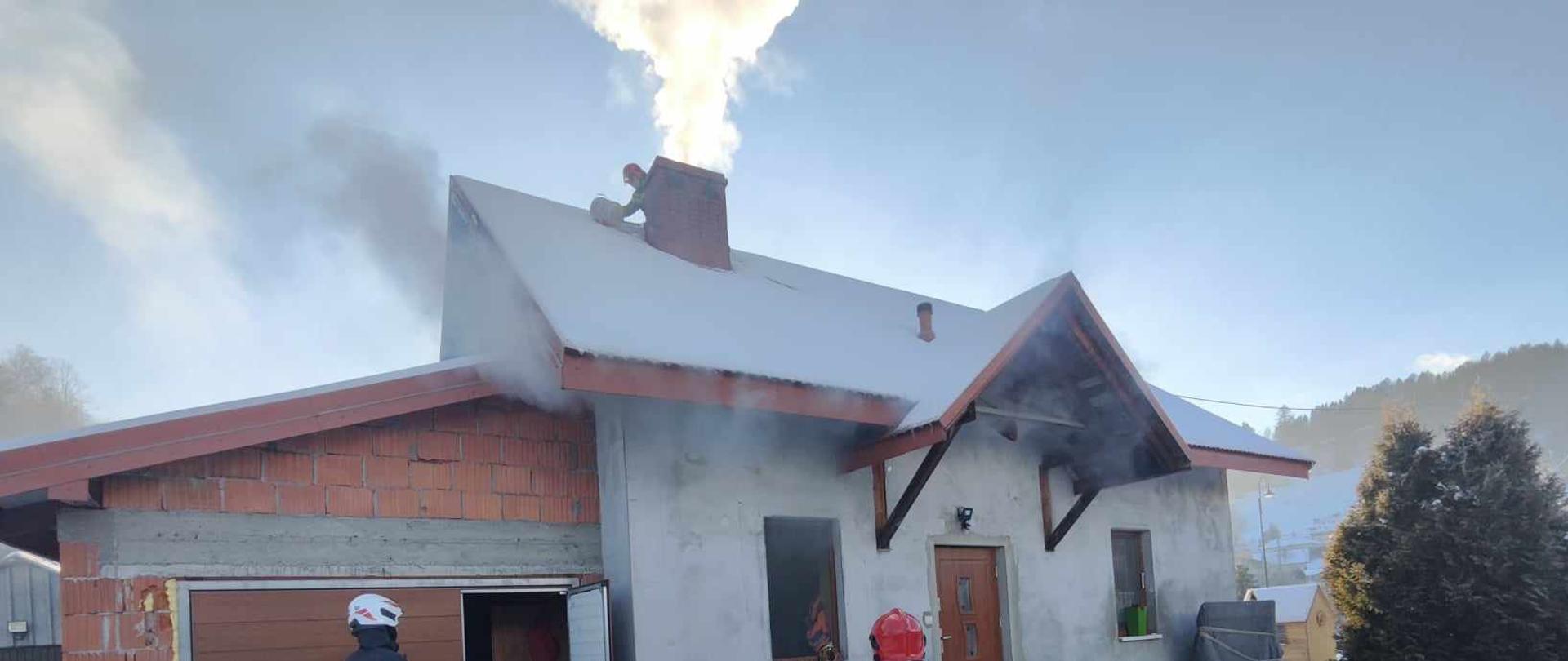 Strażacy obok budynku a na dachu strażak obok komina .