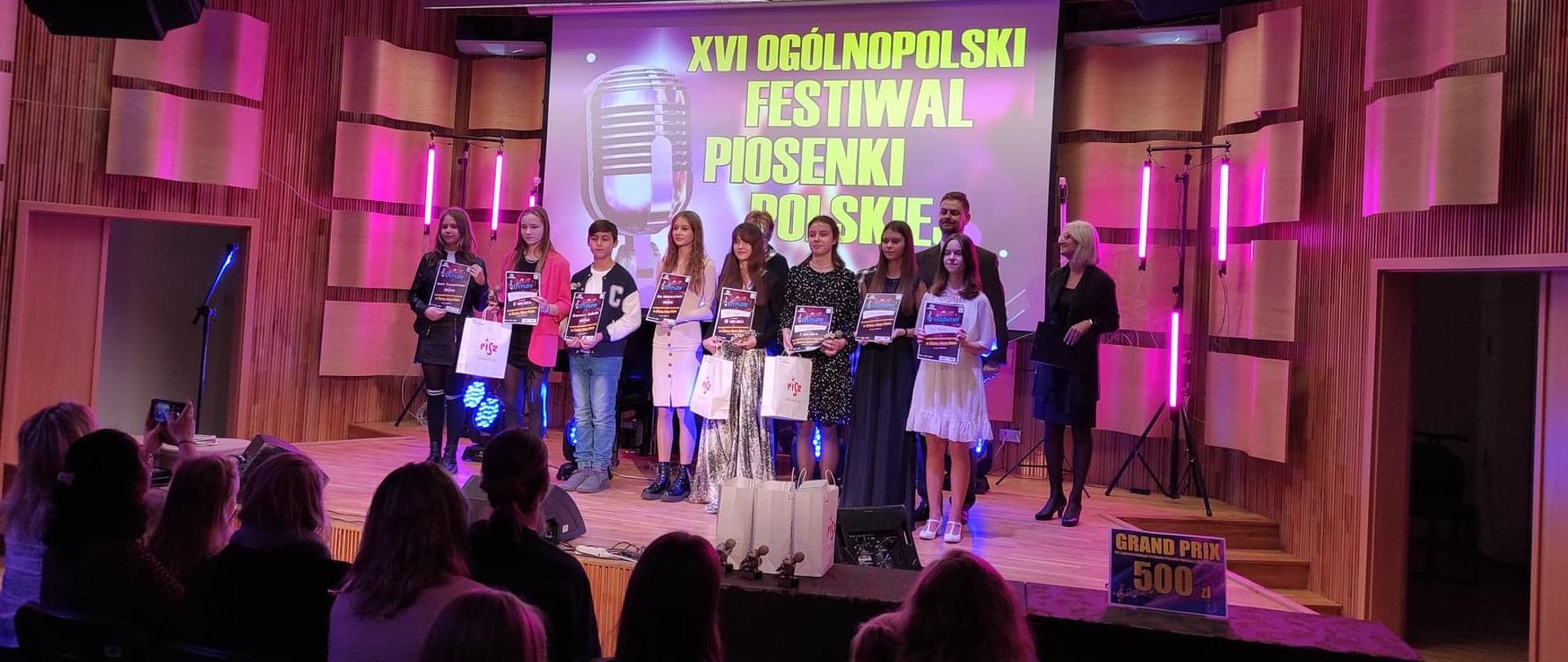 Zdjęcie laureatów konkursu na scenie OSM I st. w Piszu. W tle panele akustyczne, u dołu kontury publiczności widziane z tyłu.