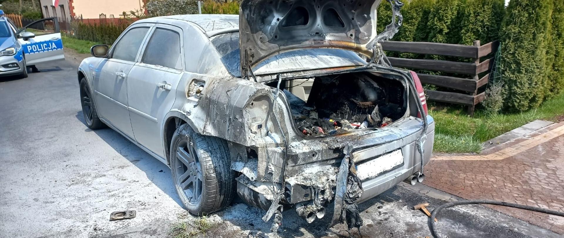 Po środku stoi samochód osobowy. Widoczne ślady po pożarze bagażnika i tyłu pojazdu. Po lewej stronie radiowóz policyjny