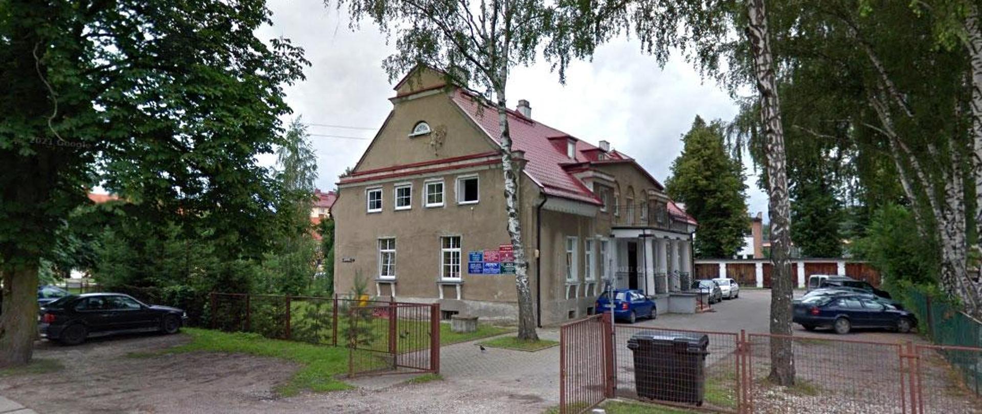 Zabytkowy budynek otoczony zielenią. Posiada charakterystyczny czerwony dach i ozdobne detale architektoniczne.