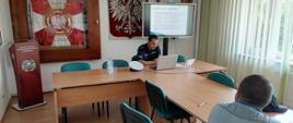 Zdjęcie w pomieszczeniu świetlicy Komendy Powiatowej PSP w Kolbuszowej. Przy stołach siedzi policjant oraz jedna osoba cywilna. Przed policjantem na stole znajdują laptop oraz czapka. Z tyłu za policjantem na monitorze wyświetlany jest tekst, na ścianie wiszą godło Polski oraz sztandar w przeszklonej gablocie. 
