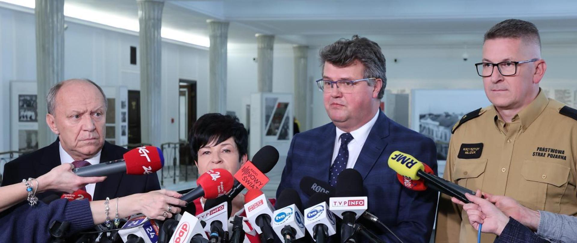 Na zdjęciu na pierwszym planie uwidoczniony jest wiceminister Maciej Wąsik, przewodniczący Parlamentarnego Zespołu Strażaków Zbigniew Chmielowiec, poseł Joanna Borowiak oraz zastępca komendanta głównego PSP nadbryg. Krzysztof Hejduk. Wiceminister wraz z towarzyszącymi mu osobami udziela wywiadu na konferencji prasowej, która odbyła się w sejmie. 