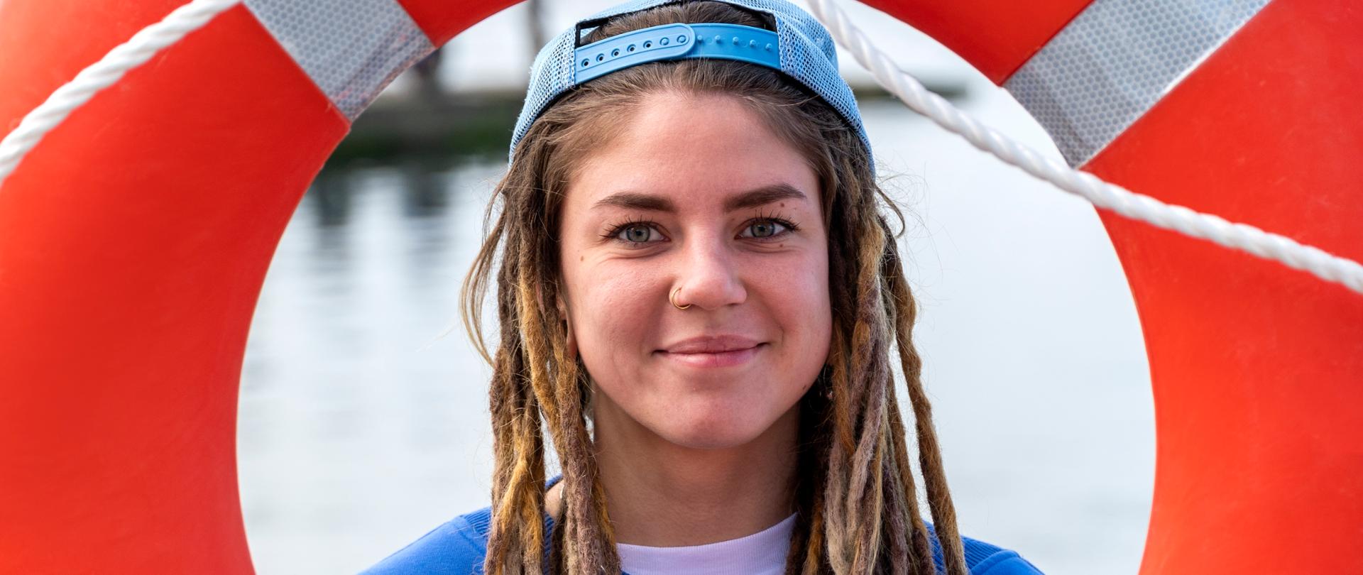 Dziewczyna w niebieskiej bluzie i czapce z daszkiem patrzy przez czerwone koło ratunkowe w stronę obiektywu.