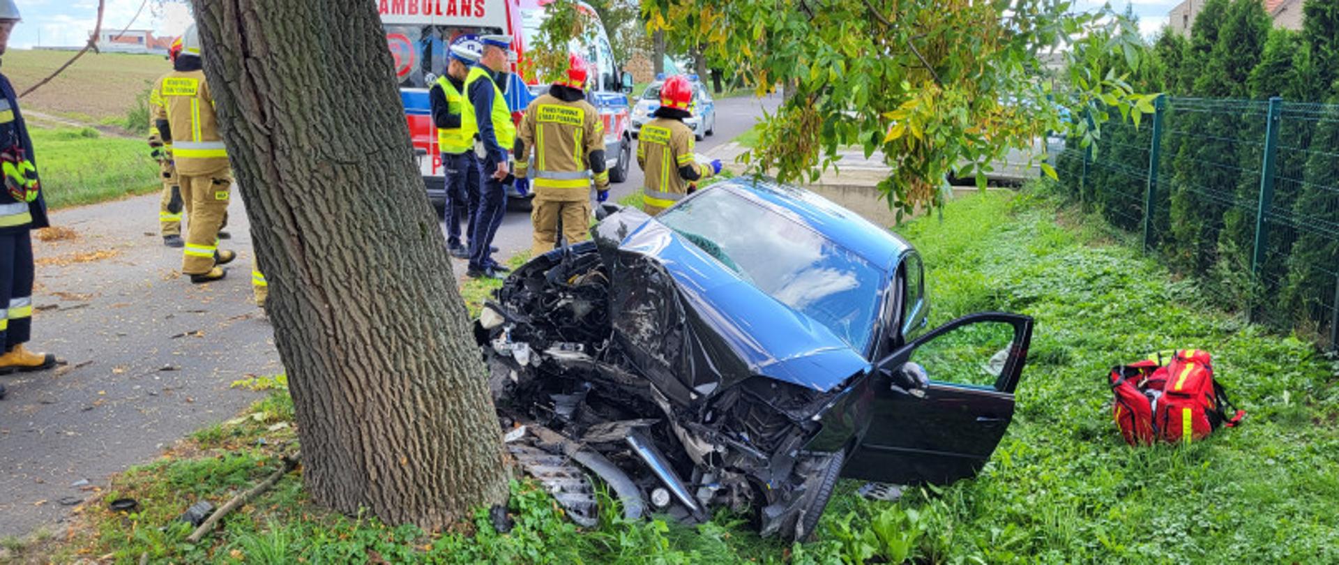 Uszkodzony samochód osobowy po uderzeniu w drzewo. W tle strażacy oraz ZRM.