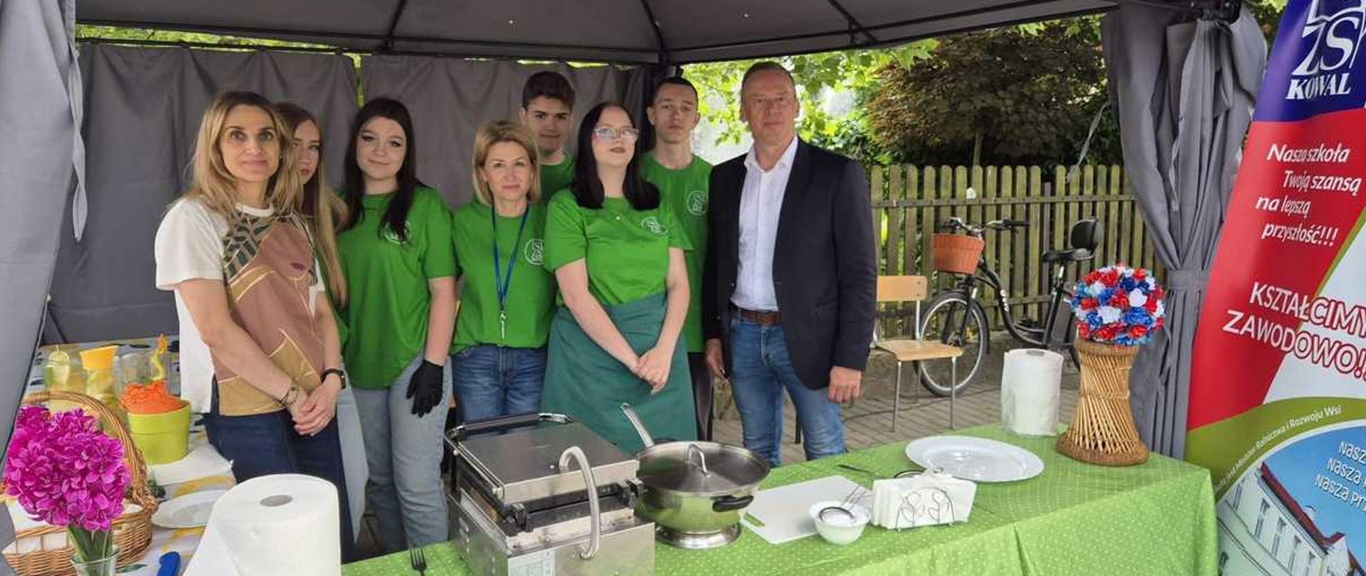 Zdjęcie przedstawia grupę siedmiu osób stojących na stoisku promocyjnym pod szarym namiotem ogrodowym. Stoisko ma charakter gastronomiczno-prezentacyjny, a w tle widać podwórko, drewniany płot i zieleń.Układ osób i ubiórOsoby stoją w dwóch rzędach za podłużnym stołem. Od lewej do prawej są to:Kobieta (pierwszy plan, lewa strona): ma długie blond włosy, jasną wzorzystą koszulkę i jeansy.Młoda kobieta (lekko schowana): ma długie, jasne włosy.Młoda kobieta: ma ciemne włosy, zieloną koszulkę i jeansy. Na dłoniach ma czarne rękawiczki jednorazowe.Kobieta w średnim wieku: ma blond włosy do ramion, zieloną koszulkę, jeansy oraz identyfikator na niebieskiej smyczy.Młody mężczyzna (w głębi): ma krótkie, ciemne włosy i zieloną koszulkę.Młoda kobieta: ma okulary, ciemne włosy i zieloną koszulkę, na którą założony ma długi, zielony fartuch kucharski.Młody mężczyzna (w głębi): ma krótkie włosy i zieloną koszulkę.Mężczyzna w średnim wieku (prawa strona): ma krótkie włosy, białą koszulę rozpiętą pod szyją, granatową marynarkę, niebieskie jeansy i brązowy pasek.Większość młodszych osób oraz jedna z kobiet nosi identyczne zielone koszulki z logo szkoły „ZS KOWAL”.Wyposażenie stoiska i otoczenieStoły i sprzęt gastronomicznyPrzed grupą ustawione są stoły przykryte jasnozielonymi obrusami w drobne, białe kropki:Główny stół: znajduje się na nim elektryczny opiekacz (kontakt-grill), metalowy garnek z pokrywką, biała deska do krojenia, miseczka, serwetki oraz ręcznik papierowy.Boczny stół (po lewej): stoi na nim wiklinowy koszyk z fioletowymi kwiatami w wazonie, żółte i pomarańczowe miski oraz akcesoria kuchenne.Dekoracja (po prawej): na rogu stołu stoi pleciony wazon z bukietem sztucznych kwiatów w barwach narodowych (biało-czerwono-niebieskie pompony).Materiały reklamowe i tłoPrawa strona: widoczny jest fragment pionowego baneru (roll-up) z fioletowym logo „ZS KOWAL”, hasłami „Nasza szkoła Twoją szansą na lepszą przyszłość!!!” i „KSZTAŁCIMY ZAWODOWO!!!” oraz zdjęciem budynku szkoły.Tło za namiotem: drewniany, brązowy płot sztachetowy, zaparkowany rower z koszykiem oraz drewniane krzesło. Nad wszystkim wiszą gałęzie drzew.
