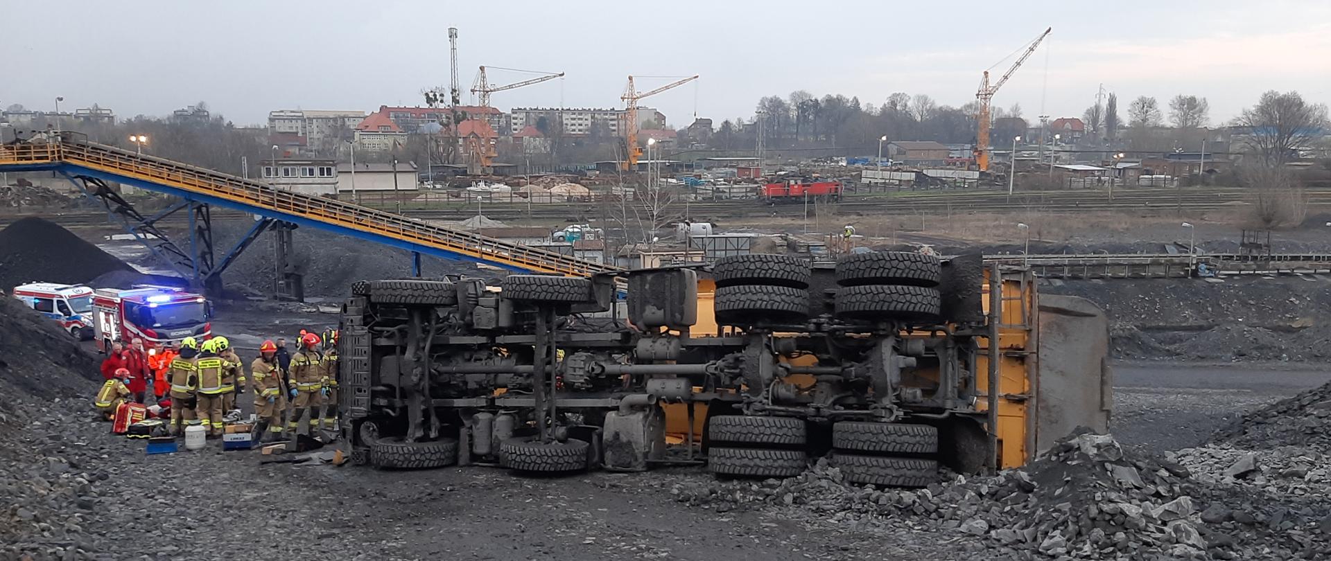 Na zdjęciu ciężarówka przewrócona na prawym boku. W tle strażacy oraz pojazdy straży pożarnej i pogotowia.