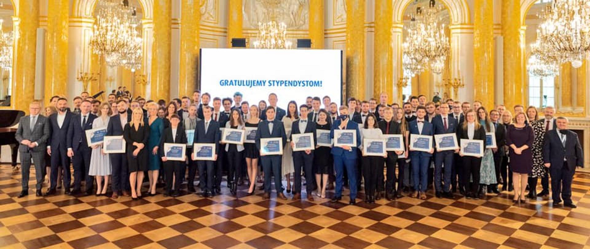 65 studentów wyróżnionych w Jubileuszowym Programie Stypendialnym 65/65, stypendyści oraz organizatorzy gali pozują do wspólnego zdjęcia we wnętrzach Zamku Królewskiego w Warszawie.