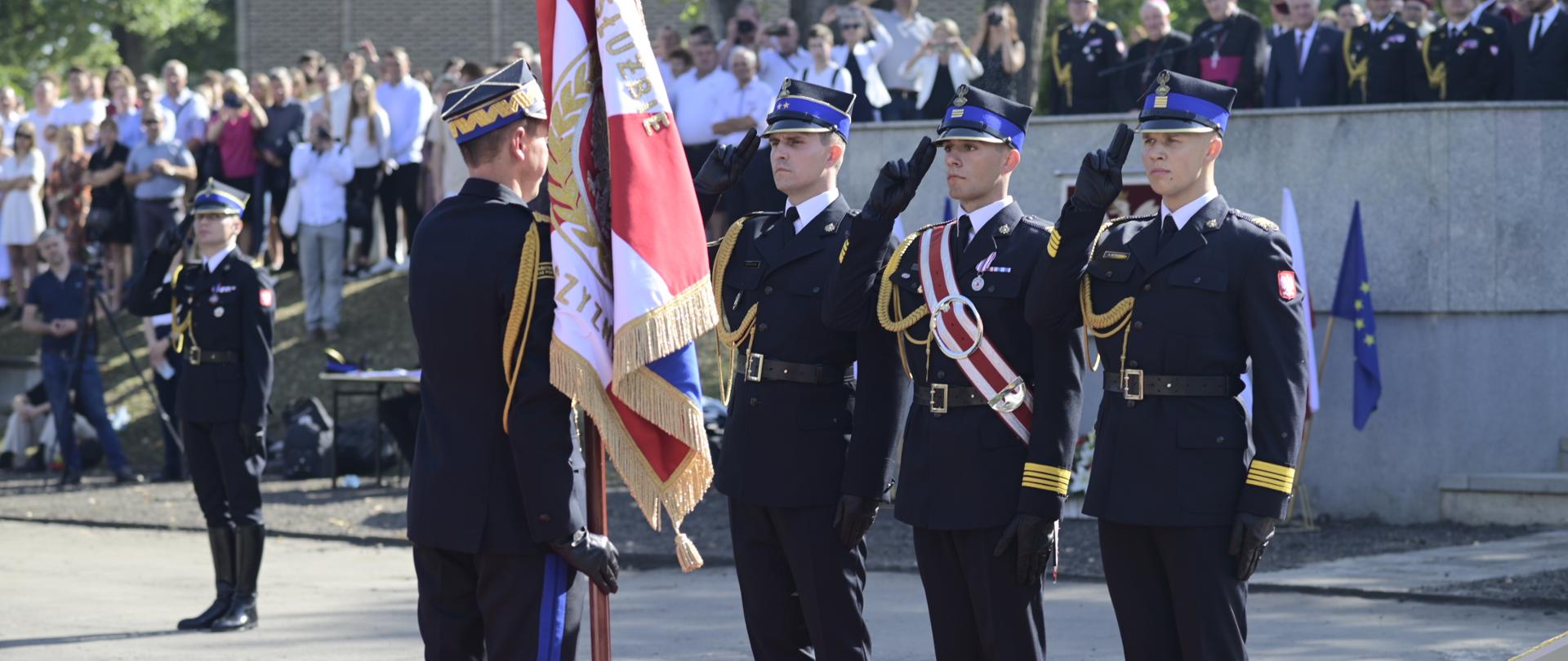 Na placu w ostrym słońcu stoi mężczyzna w czarnym strażackim galowym mundurze, trzyma sztandar, przed nim stoi trzech strażaków i salutuje.