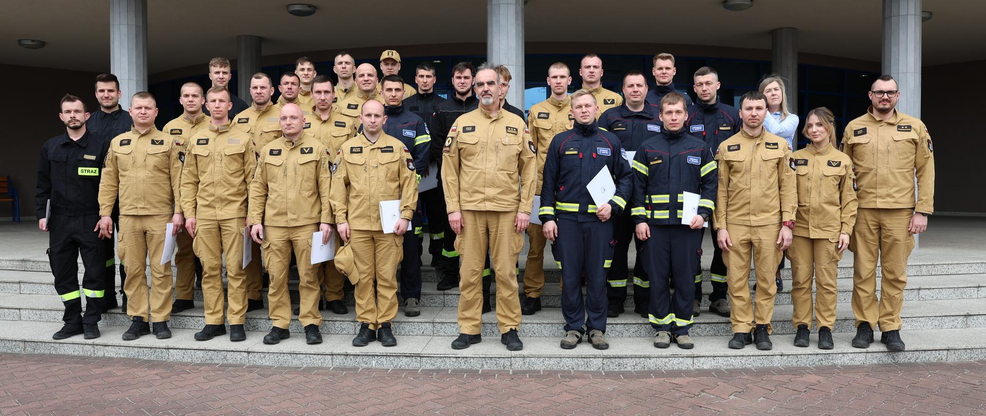 Pamiątkowe zdjęcie grupowe funkcjonariuszy PSP będących uczestnikami szkolenia w zakresie ratownictwa chemicznego i ekologicznego (26 osób) w towarzystwie Zastępcy Komendanta Centralnej Szkoły PSP, kobiety oraz trojga funkcjonariuszy kadry dydaktycznej CS PSP